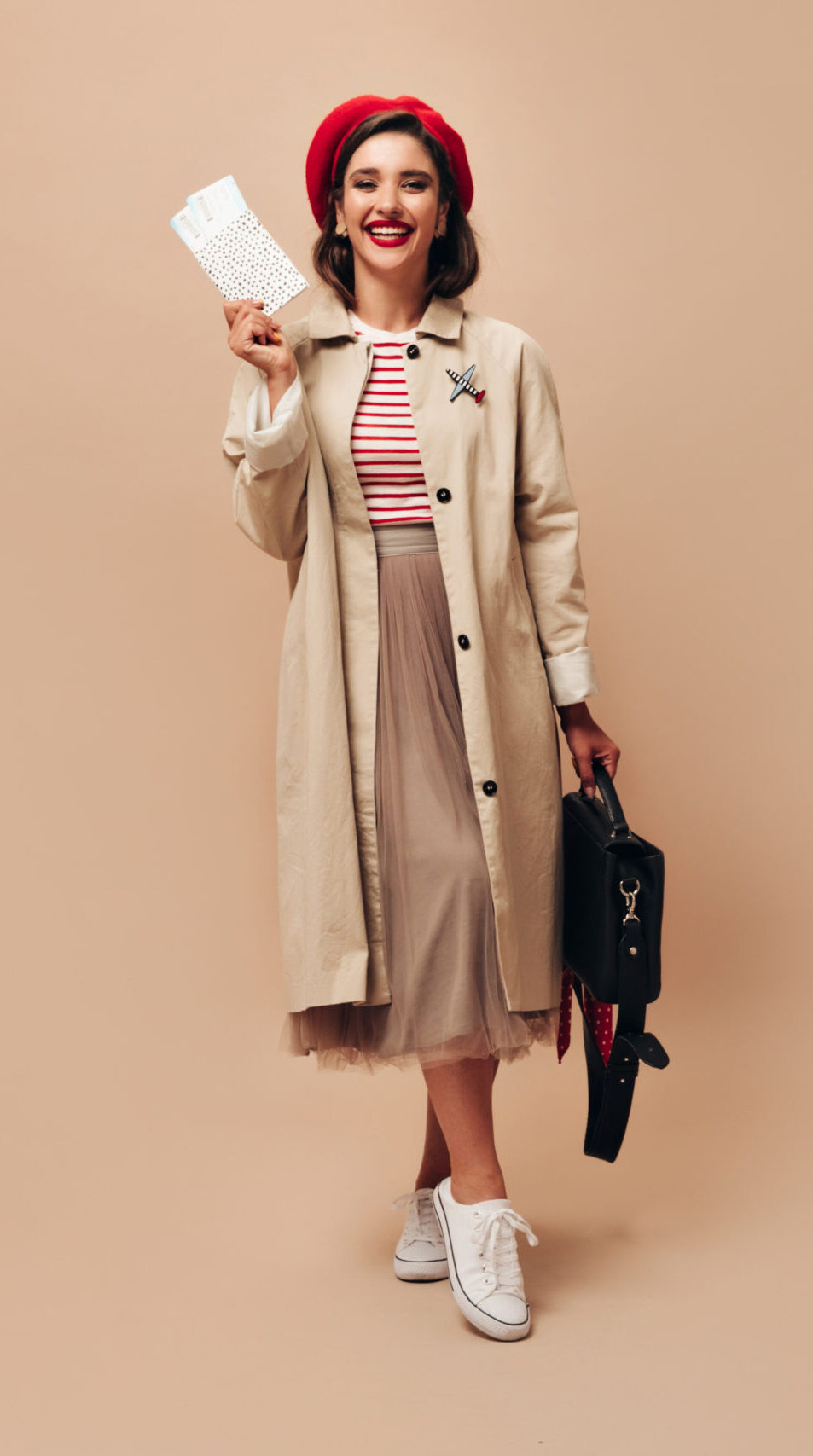 Happy Lady on red beret and trench poses with tickets on beige background. Joyful young woman in long stylish skirt and autumn coat looks at camera..